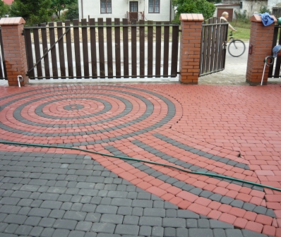 Ułożona kostka brukowa w kolorach czerwieni i szarości tworząca wzór koła i promieni na podjeździe przed bramą z drewnianymi sztachetami i ceglanymi słupkami. W tle widoczny dom i osoba na rowerze.