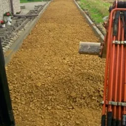 Utwardzanie drogi gruzem, widok z kabiny koparki, zarys budynków w tle.