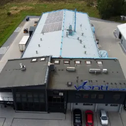 Widok z góry na budynek firmy Venti - Lustra Szkło z zainstalowanymi panelami słonecznymi na dachu, widoczny również fragment parkingu z samochodami i teren zielony.
