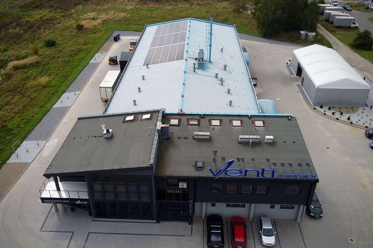 Widok z góry na budynek firmy Venti - Lustra Szkło z zainstalowanymi panelami słonecznymi na dachu, widoczny również fragment parkingu z samochodami i teren zielony.
