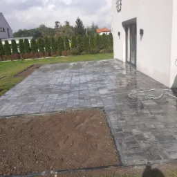 Świeżo ułożona, mokra kostka brukowa w Bielsko-Białej, przylegająca do nowoczesnego domu z dużym oknem tarasowym, obok fragment ziemi przygotowany pod dalsze prace.