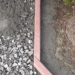 Przygotowanie podłoża pod ścieżkę z obrzeżami z czerwonej kostki, widoczne kamienie, piasek i wyrównana ziemia wzdłuż krawężnika.