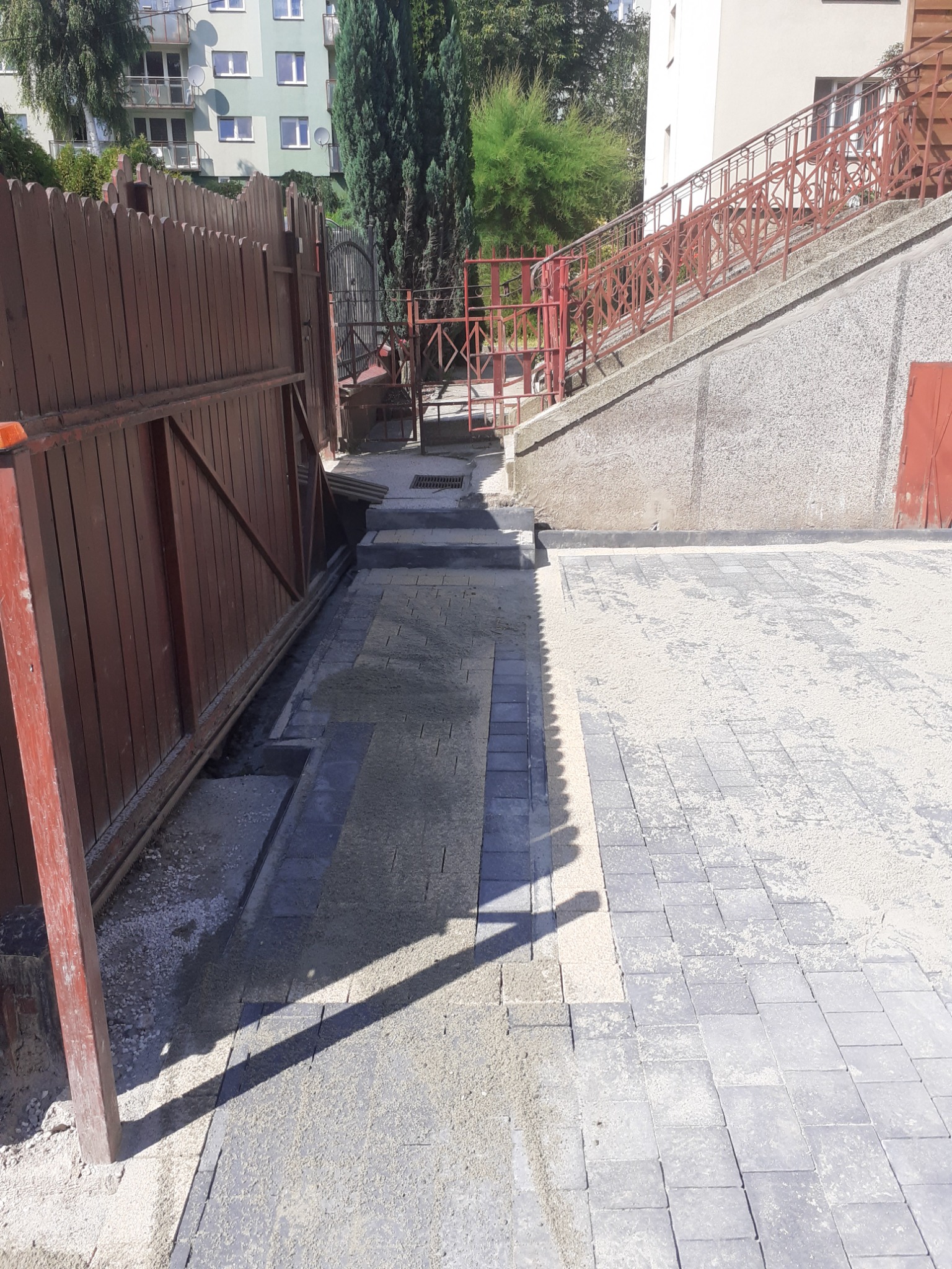 Fragment nowo ułożonej, szarej kostki brukowej na posesji z drewnianą bramą w tle, widoczny etap zasypywania piaskiem i częściowo ułożone korytko odwadniające.