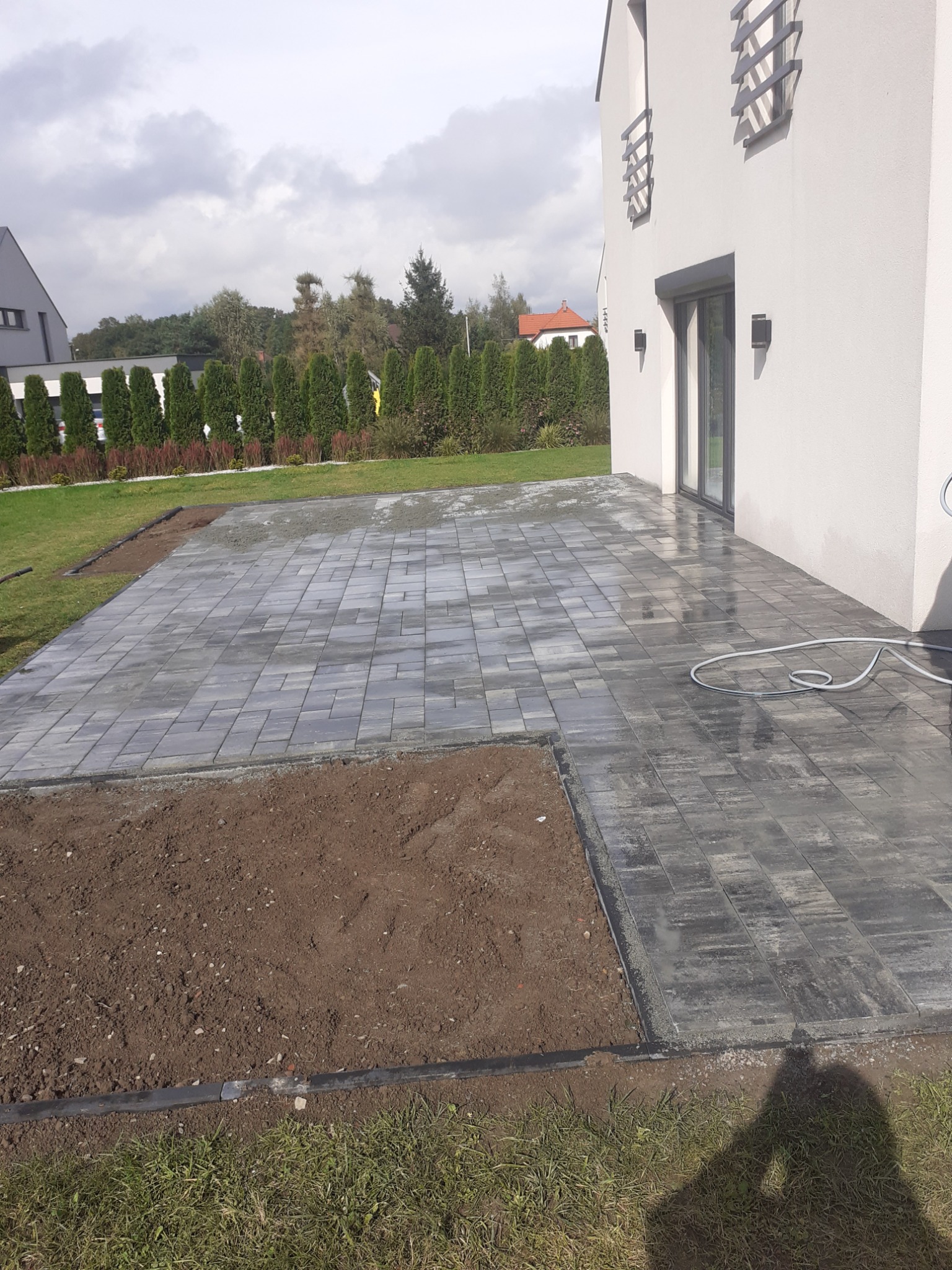 Świeżo ułożona, mokra kostka brukowa w Bielsko-Białej, przylegająca do nowoczesnego domu z dużym oknem tarasowym, obok fragment ziemi przygotowany pod dalsze prace.