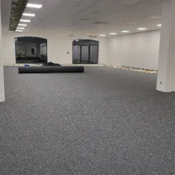 Wnętrze biurowe po położeniu szarej wykładziny dywanowej, widoczne kolumny konstrukcyjne, przygotowane rolki wykładziny i drabina w tle.