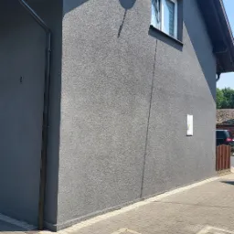 Ściana domu jednorodzinnego z szarą elewacją o strukturze baranka, widoczny fragment okna z białymi ramami, antena satelitarna na elewacji, rynna, kostka brukowa na chodniku.