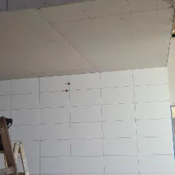 Montaż płyt kartonowo-gipsowych na ścianie i suficie, widoczna drewniana drabina i surowe wykończenie poddasza z izolacją.