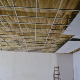 DAM-BUD Firma Wykonczeniowo-remontowa - Szkielet sufitu podwieszanego z widoczną izolacją termiczną, drabina stojąca w pustym pomieszczeniu, kilka płyt sufitowych już zamontowanych.