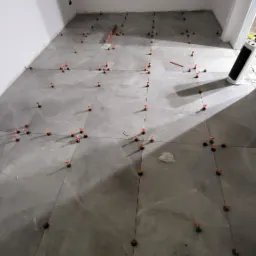 Układanie szarych płytek podłogowych w pomieszczeniu z oknem, widoczne pomarańczowe kliny poziomujące i krzyżyki dystansowe, oświetlenie sufitowe tworzy cień na podłodze.