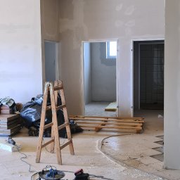DAM-BUD Firma Wykonczeniowo-remontowa - Wnętrze w trakcie prac remontowych: ocieplony żółtą wełną mineralną sufit, ściany częściowo wykończone gładzią, drewniana drabina, rozsypany gruz na podłodze z płytek i zwinięty przedłużacz...
