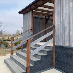 ELBOWOOD Andzej Borkowski - Drewniane schody wejściowe z balustradą, prowadzące do zadaszonego wejścia budynku. Elementy konstrukcyjne w odcieniach szarości i brązu. Widok z dołu, lekko z boku.