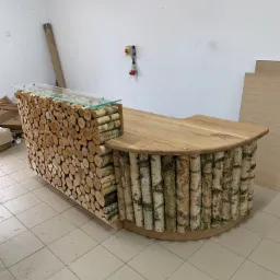 Recepcja wykonana z drewna, oklejona okrąglakami z brzozy, blat z jasnego drewna, nadstawka z szybą, surowy stan wykończenia wnętrza.
