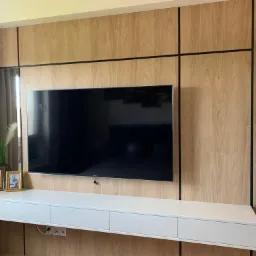 Ściana z paneli drewnopodobnych z czarnymi listwami tworzącymi geometryczny wzór, wiszący telewizor, długa, biała szafka RTV z szufladami, dekoracje w postaci ramek i roślin w doniczce.