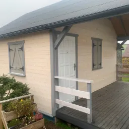 Drewniany domek letniskowy z szarymi okiennicami i zadaszonym tarasem, widok z boku, drewniana donica z kwiatami przed domkiem.