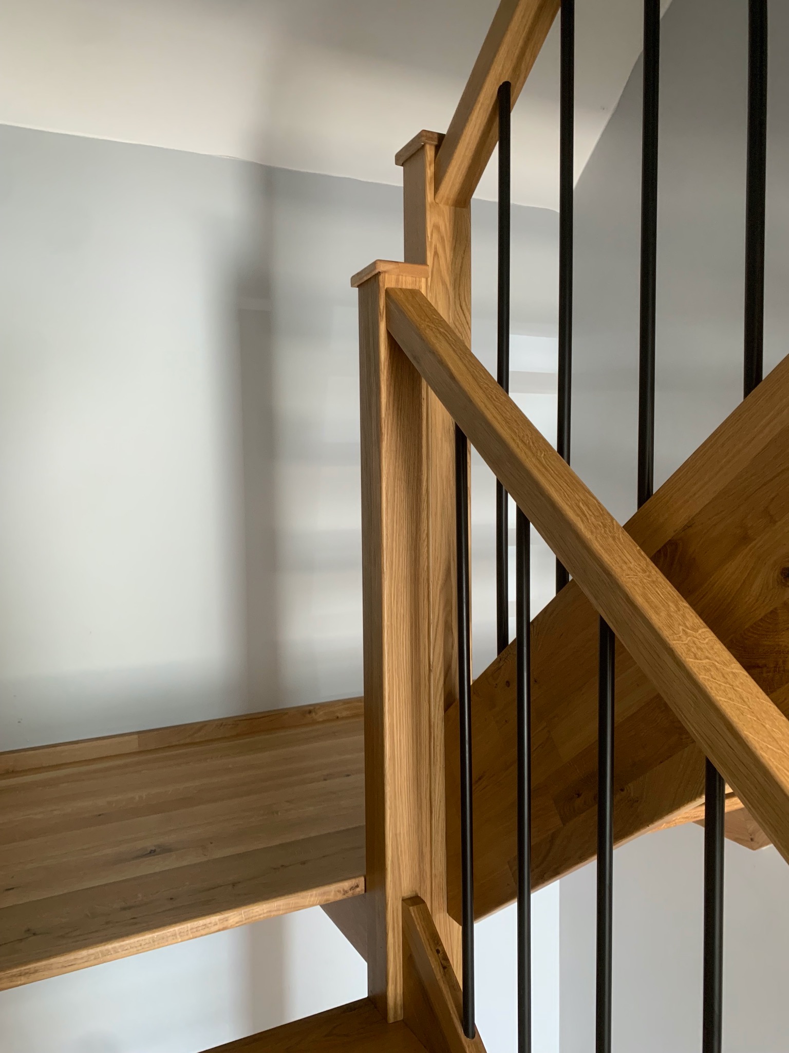 Fragment drewnianych schodów z czarnymi, metalowymi balustradami na tle jasnej ściany. Widoczne detale wykończenia drewna i nowoczesny design.