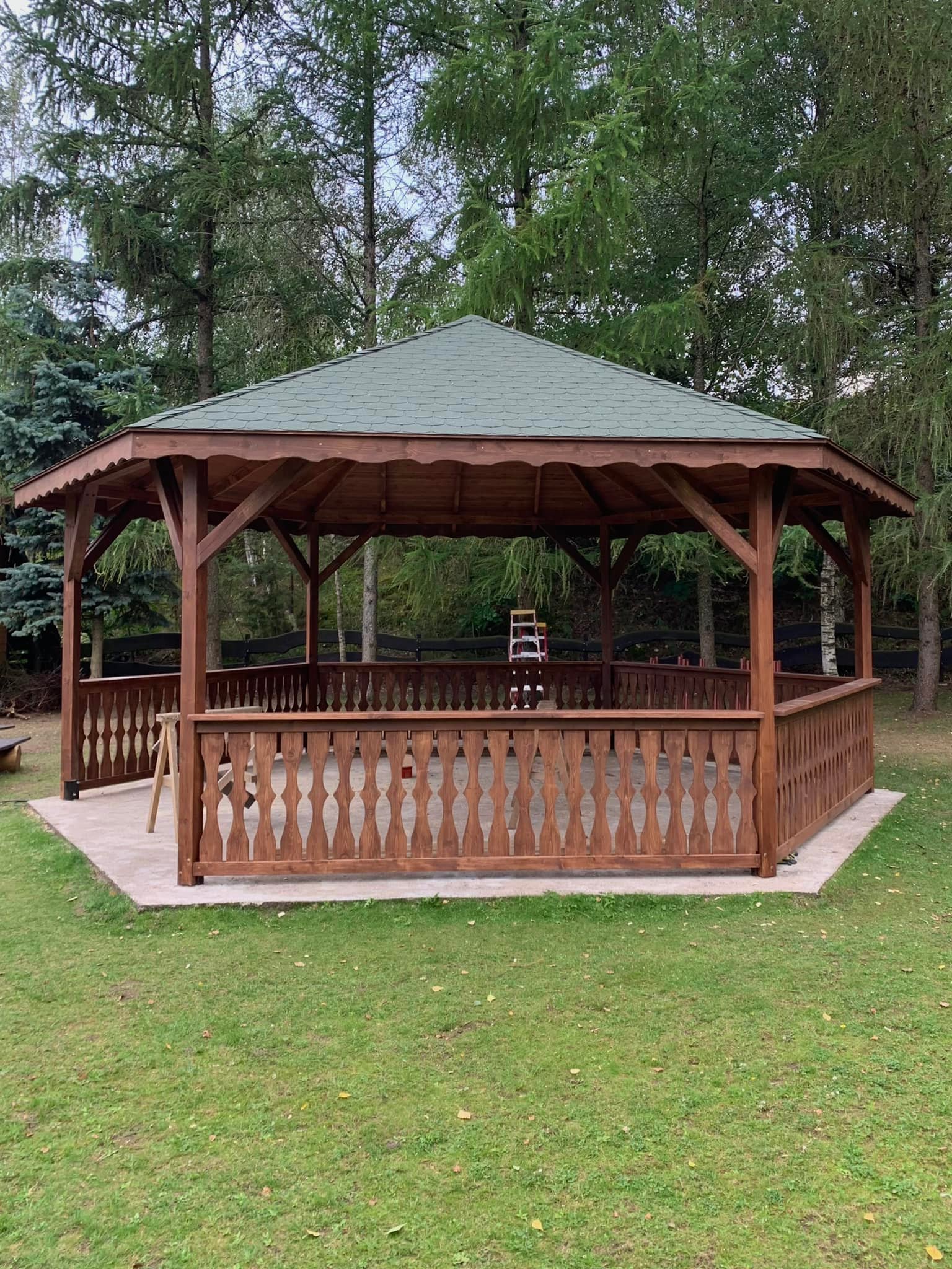 Drewniana altana ogrodowa z zielonym dachem, otoczona drzewami, z widocznymi detalami wykończenia i ażurowymi barierkami. Na trawie leżą opadłe liście.