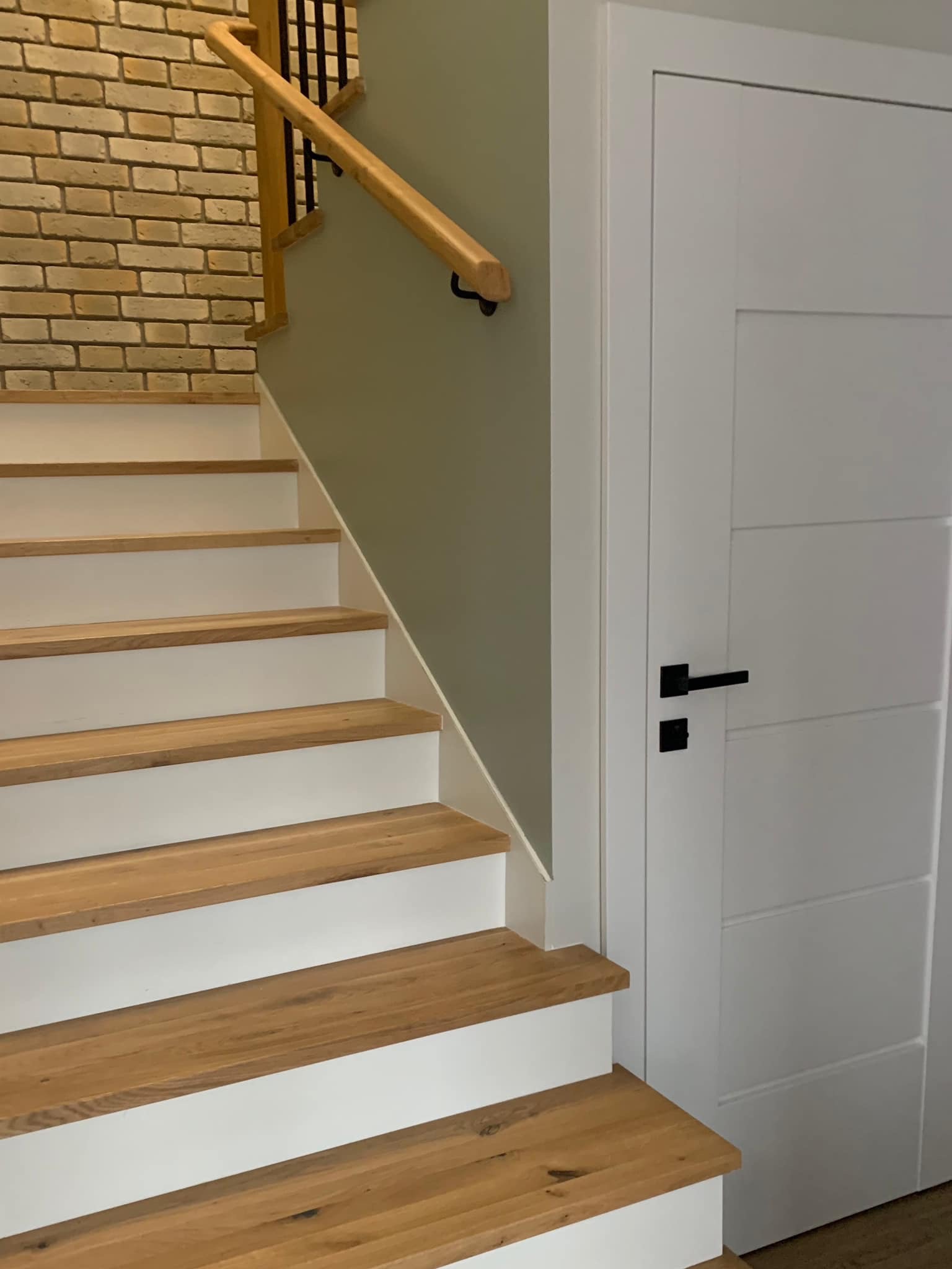 Drewniane schody z białymi podstopnicami i jasną cegłą na ścianie, obok białe drzwi z czarną klamką. Ujęcie z dołu, widoczne stopnie i fragment ściany.