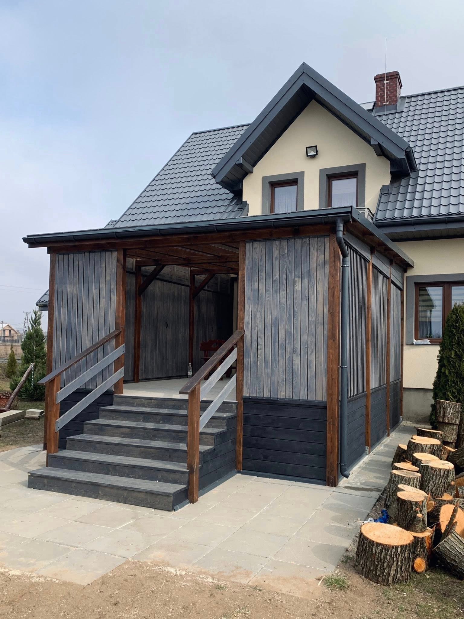 Zadaszone wejście do domu z drewnianymi schodami i ścianami w odcieniach szarości i brązu. Na pierwszym planie ułożone pniaki drewna.