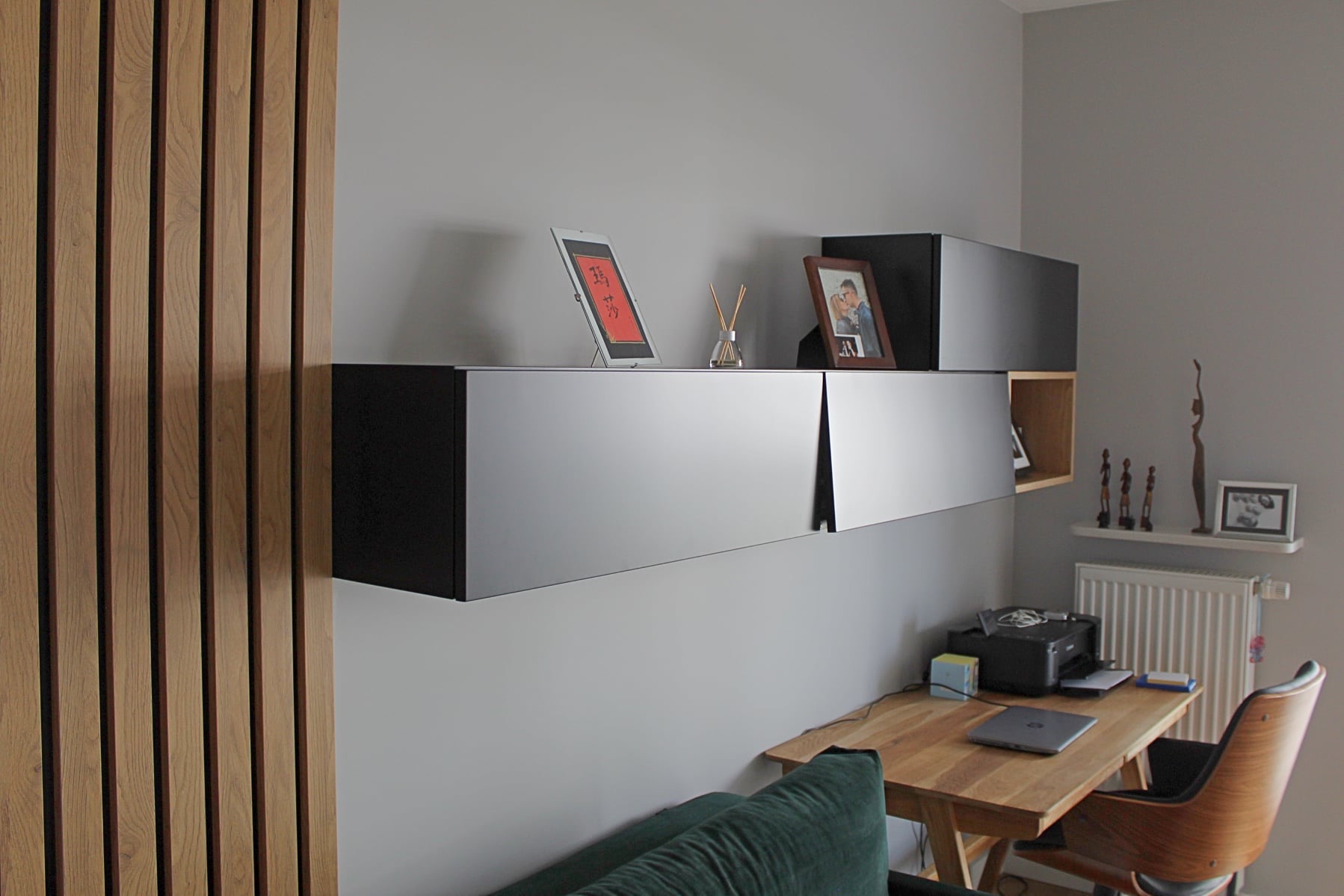 Nowoczesna szafka wisząca w salonie, z elementami drewnianej boazerii na ścianie. Minimalistyczny design, szare ściany, drewniany blat biurka i dekoracje.