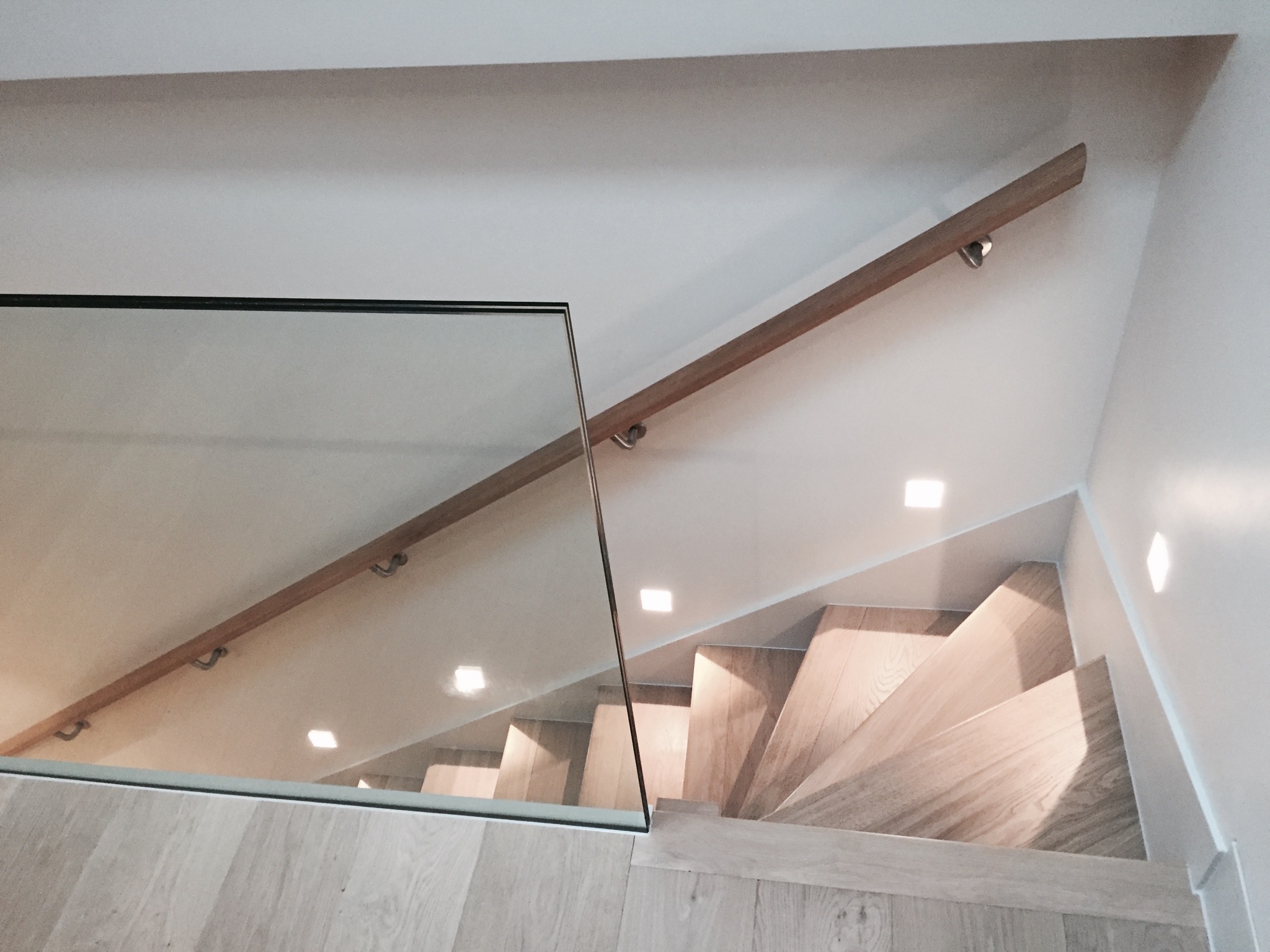 Nowoczesne schody z drewnianymi stopniami, szklaną balustradą i oświetleniem punktowym w ścianie i nad biegiem schodów.