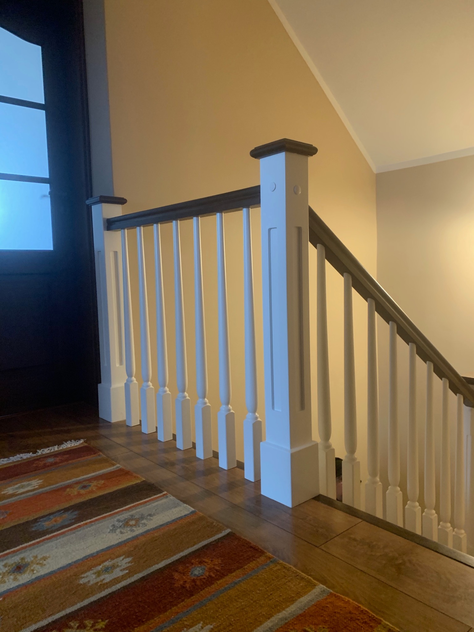 Drewniane schody z białymi balustradami i ciemnym poręczą, widok z dołu, dywan w etniczne wzory na drewnianej podłodze, drzwi z szybą w tle.