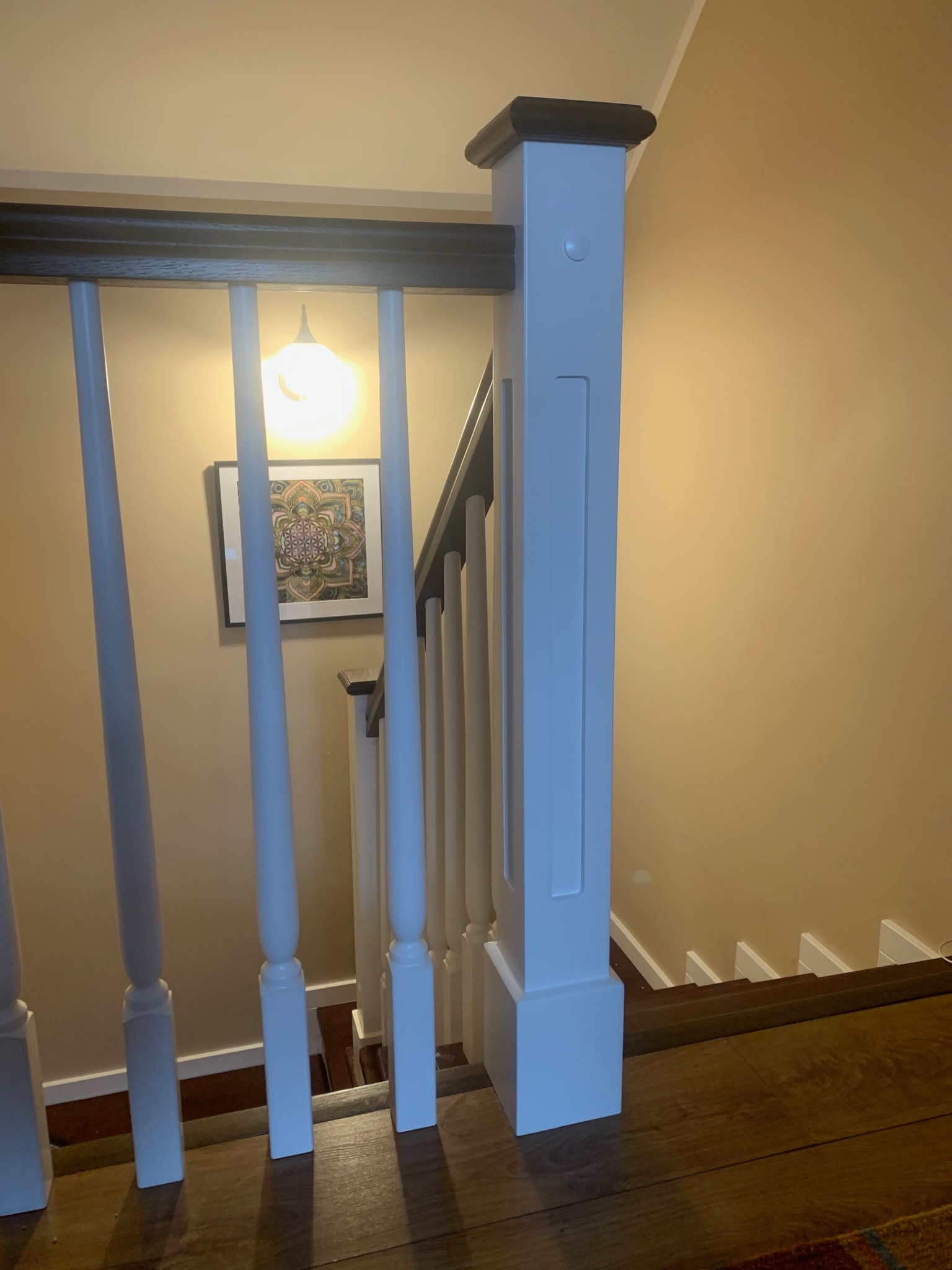 Fragment drewnianych schodów z białymi balustradami i ciemnobrązową poręczą, widok z góry na dół, na ścianie wisi obrazek w ramce.