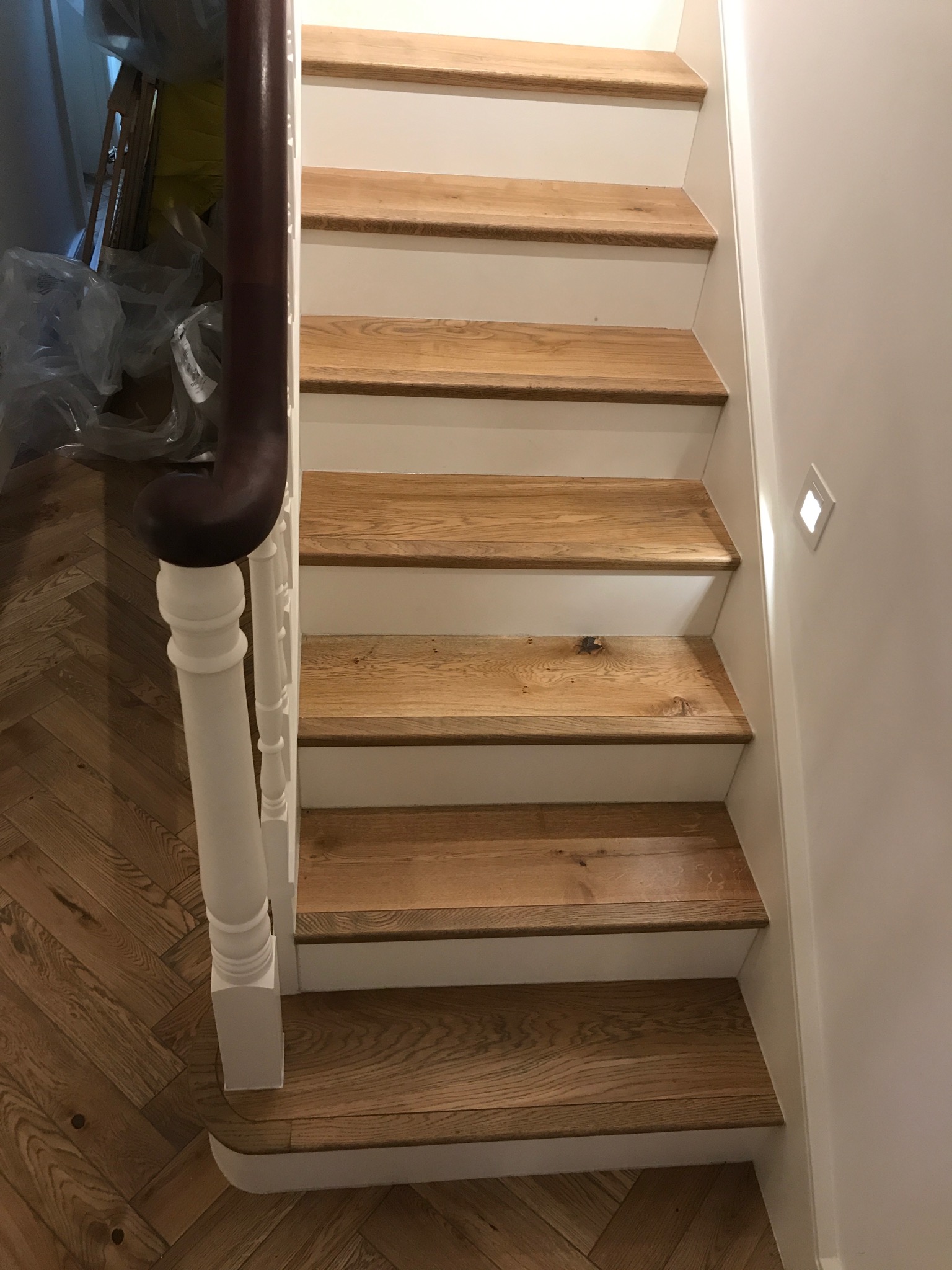 Bieg schodów z drewnianymi stopniami i białymi podstopnicami, widoczna drewniana podłoga ułożona w jodełkę oraz fragment białej balustrady z ciemnobrązowym poręczą.