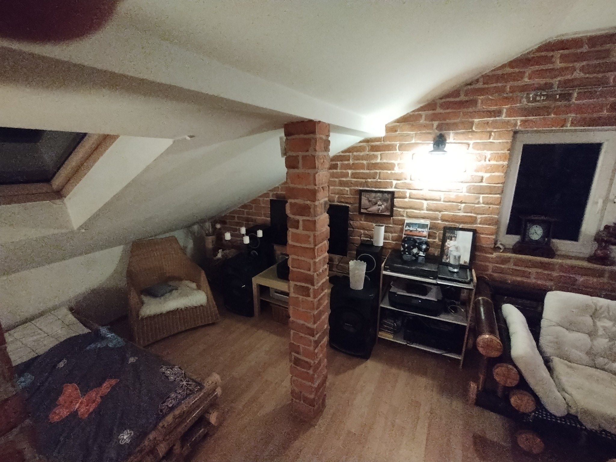 Poddasze mieszkalne z ceglaną ścianą, drewnianą podłogą i meblami w stylu rustykalnym, oświetlone lampą ścienną i światłem z okna dachowego.