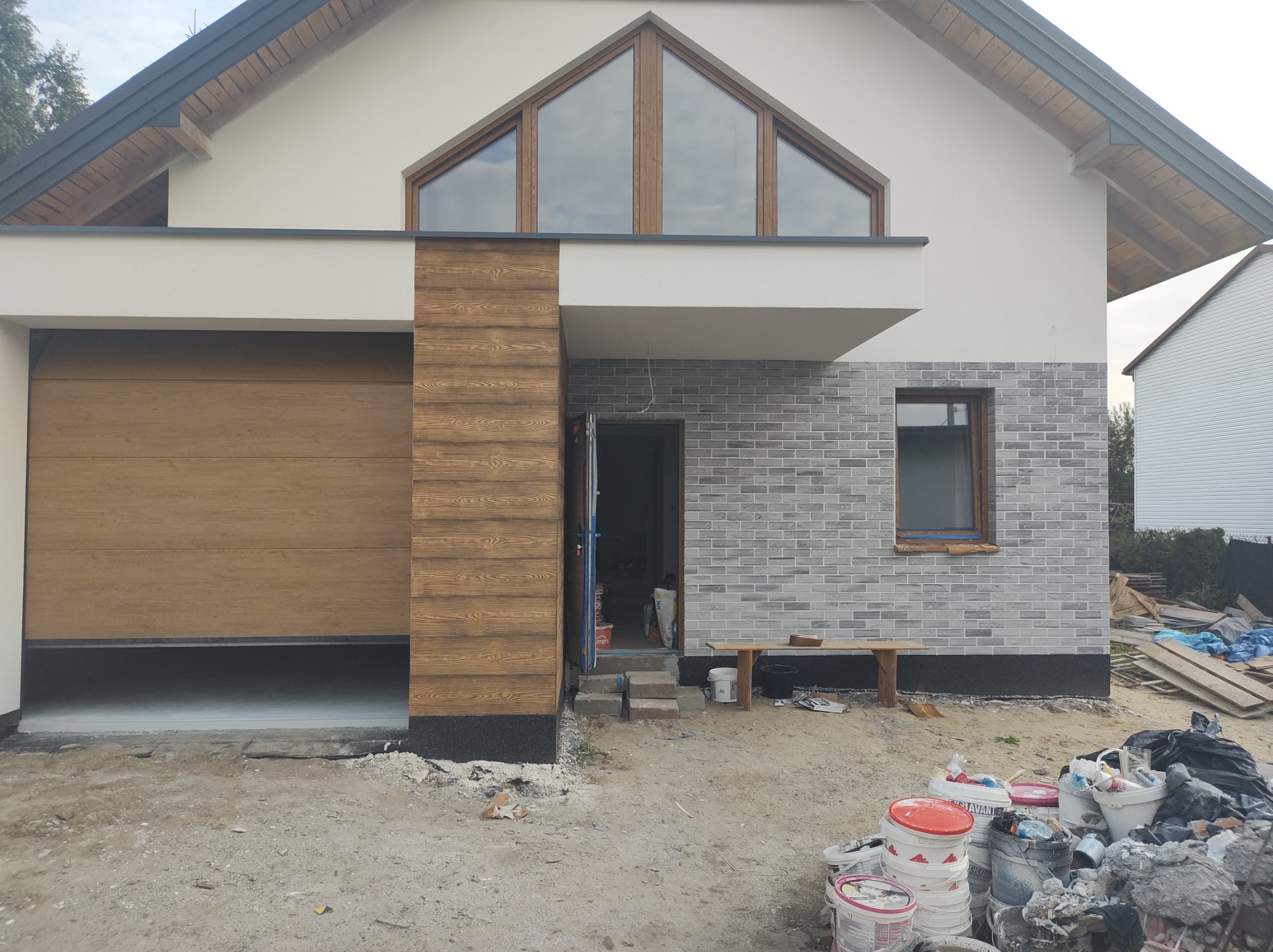 Nowoczesny dom jednorodzinny z garażem i elewacją w trakcie wykończenia: fragment ściany z szarej cegły klinkierowej, drewniana okładzina oraz otwarta brama garażowa. W tle widoczne ślady prac...