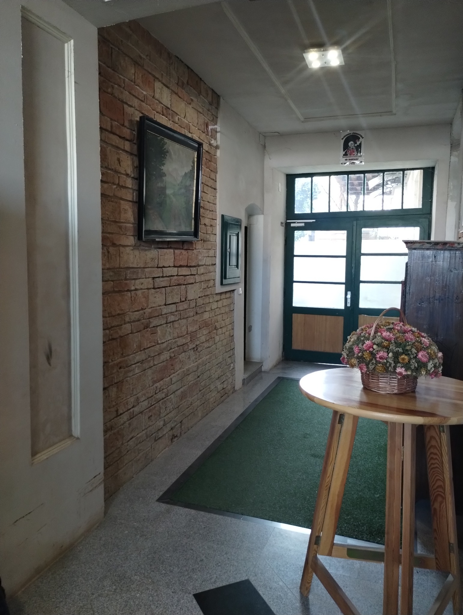 Korytarz z ceglaną ścianą, obrazem w czarnej ramie, drewnianym stolikiem z bukietem kwiatów i zielonym dywanikiem przy wejściu.