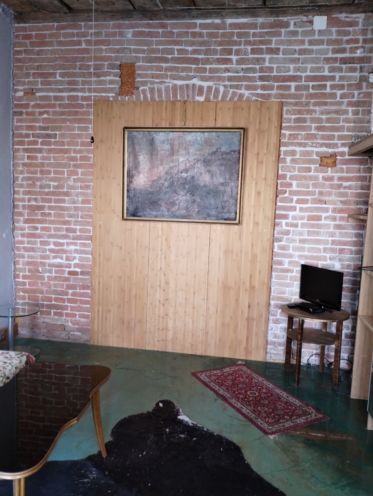 Brązowa ceglana ściana z drewnianą boazerią za obrazem w złotej ramie; podłoga w odcieniach zieleni z dywanem i czarną skórą.