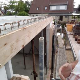 Staska Bau Rem - Wylewka betonowa stropu z widocznym zbrojeniem, szalunkiem drewnianym i podporami stalowymi, na tle domu jednorodzinnego.