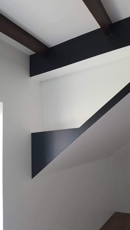 Minimalistyczne wnętrze z elementami dekoracyjnymi w kolorze ciemnego brązu i czerni, kontrastujące z bielą ścian i sufitu. Widoczne fragmenty belek stropowych i geometryczna forma schodów.