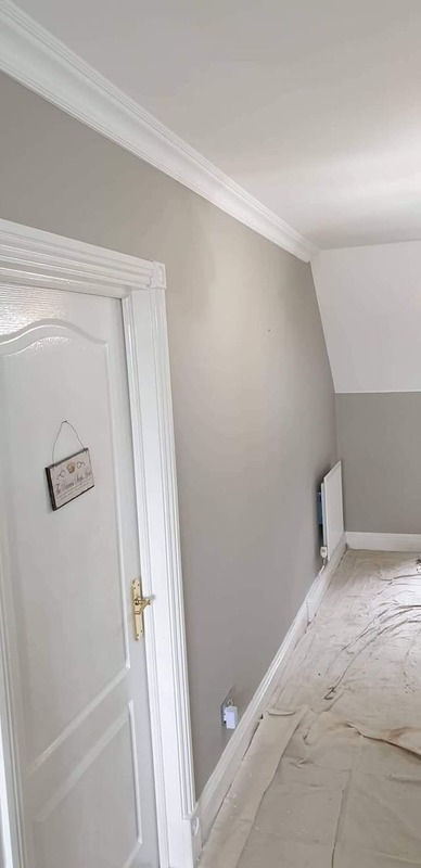 Świeżo pomalowane wnętrze pokoju w odcieniach szarości z białymi listwami przypodłogowymi i sufitowymi, zabezpieczone folią malarską, widoczna biała stolarka drzwiowa z dekoracyjnymi frezami...