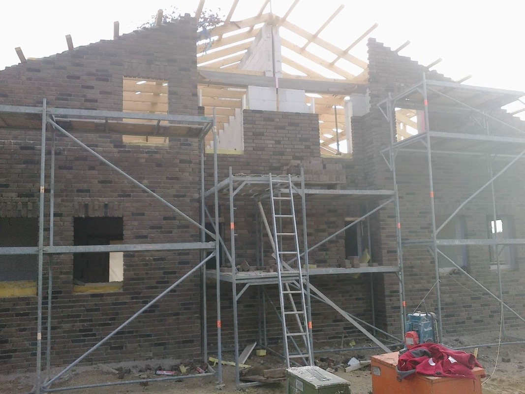 Budowa domu jednorodzinnego w trakcie murowania ścian z cegły klinkierowej, widoczne rusztowania i konstrukcja dachu.