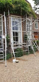 Rusztowanie rozstawione przed budynkiem z cegły, przygotowania do prac remontowych przy oknach, widoczne pojemniki z farbą.