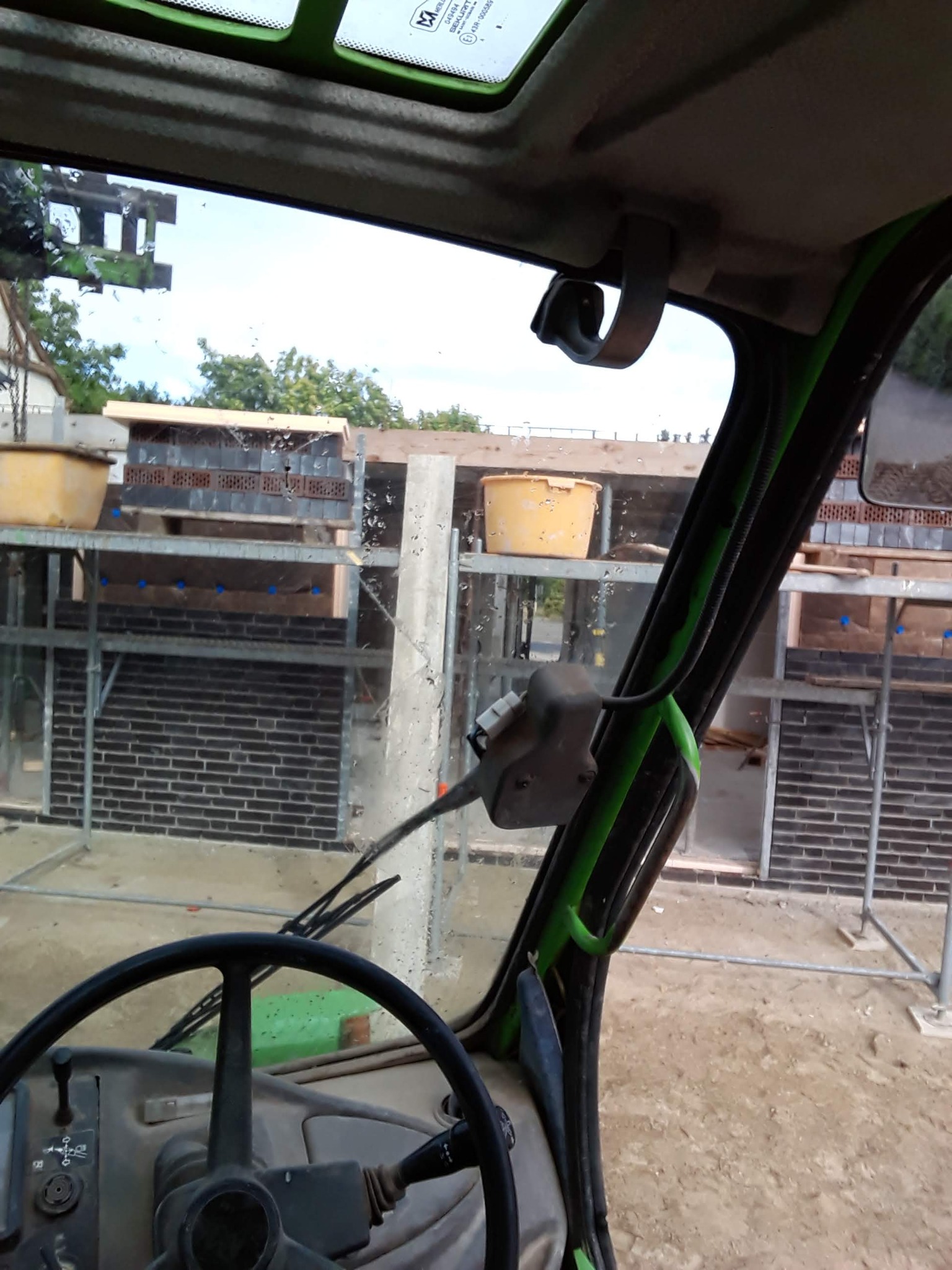 Widok z kabiny maszyny budowlanej na rusztowanie z ceglaną ścianą w trakcie budowy, z żółtymi wiadrami na górze.