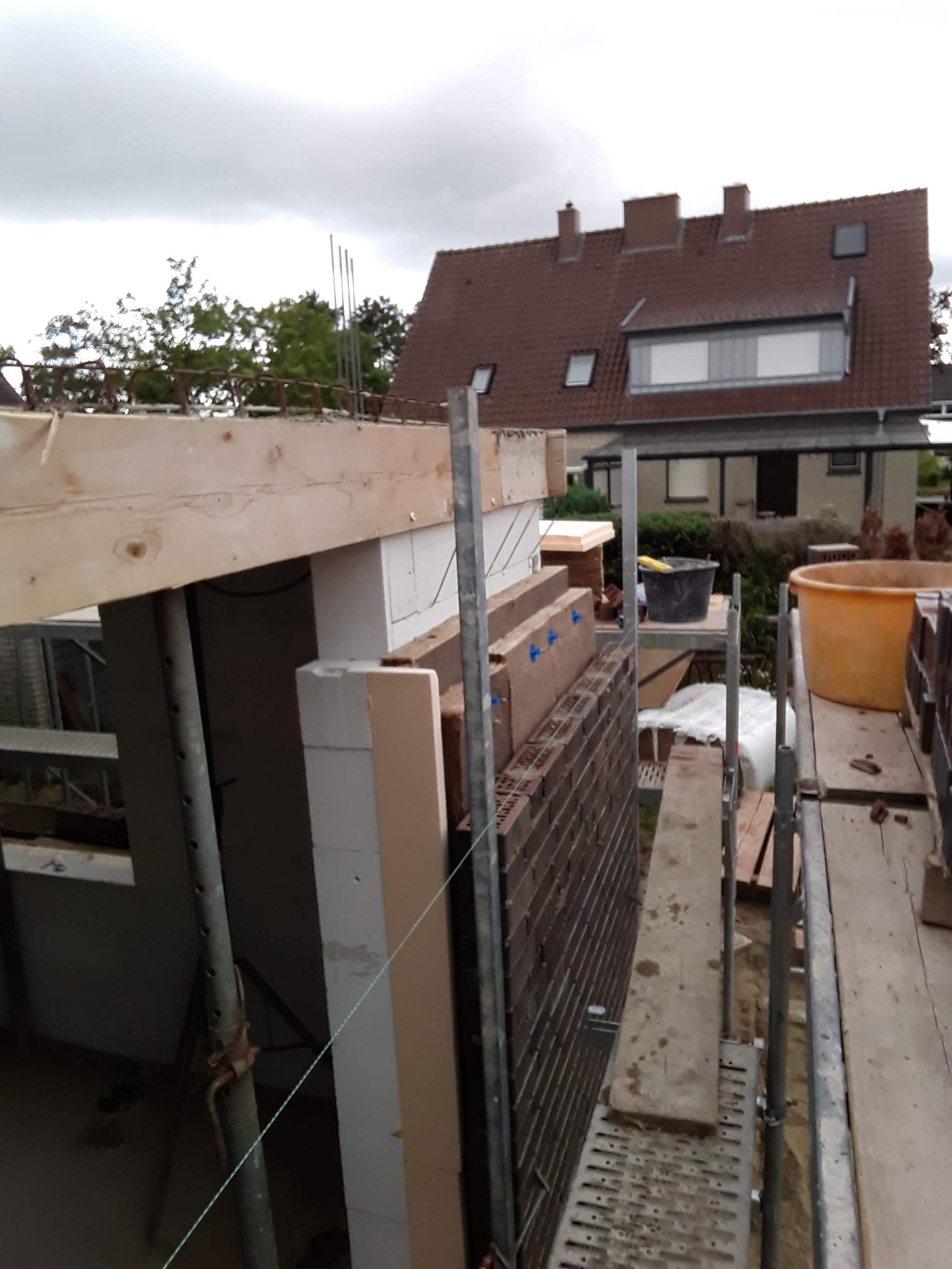 Budowa domu w trakcie: widok z rusztowania na wznoszone ściany z cegły i bloczków, elementy konstrukcji drewnianej i stalowe podpory, w tle dom z czerwoną dachówką.
