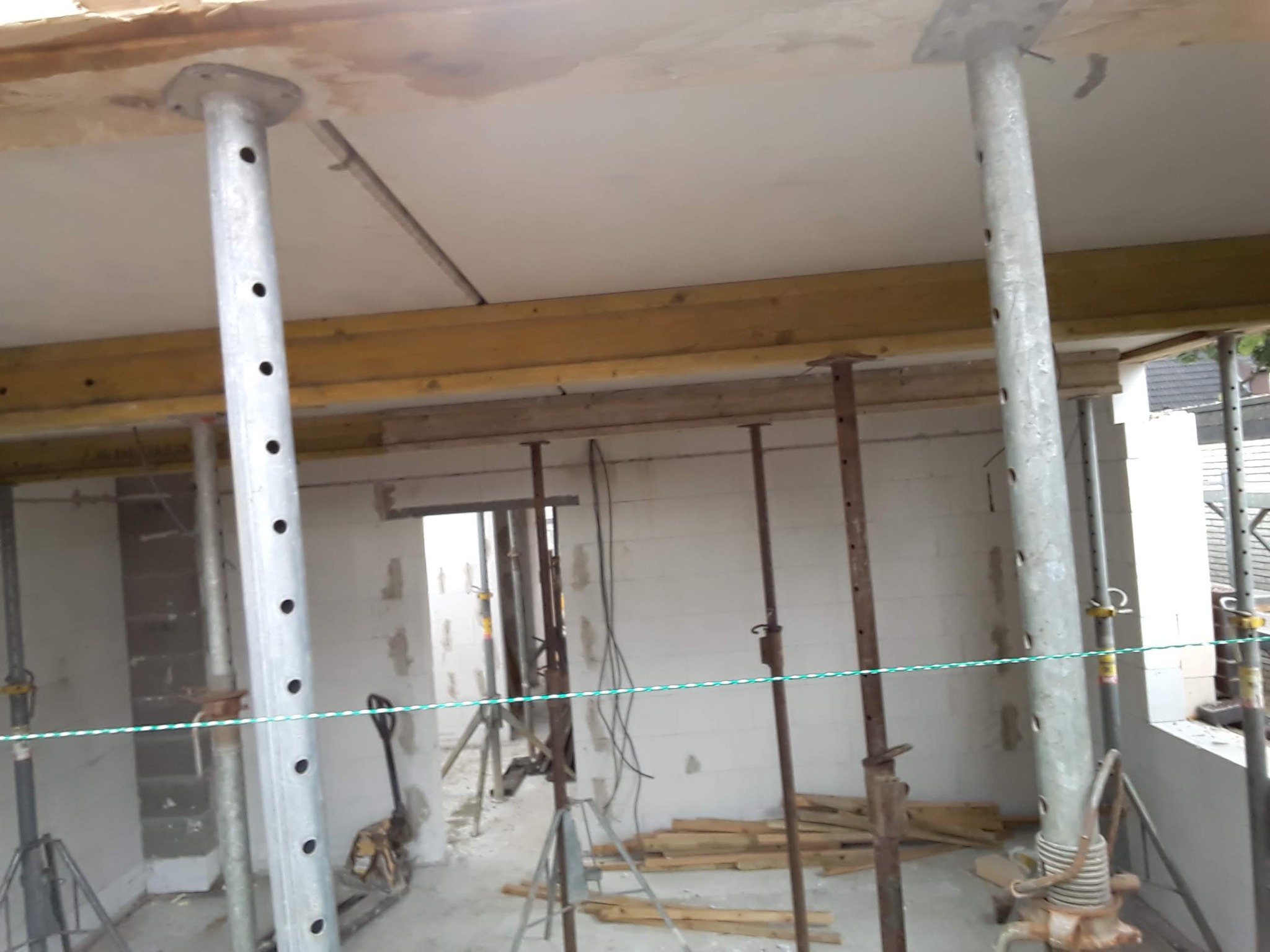 Wnętrze w trakcie budowy, widoczne podpory stropowe, belki drewniane i ściany z bloczków. Na podłodze leżą deski, a w tle widać narzędzia i kolejne pomieszczenie.