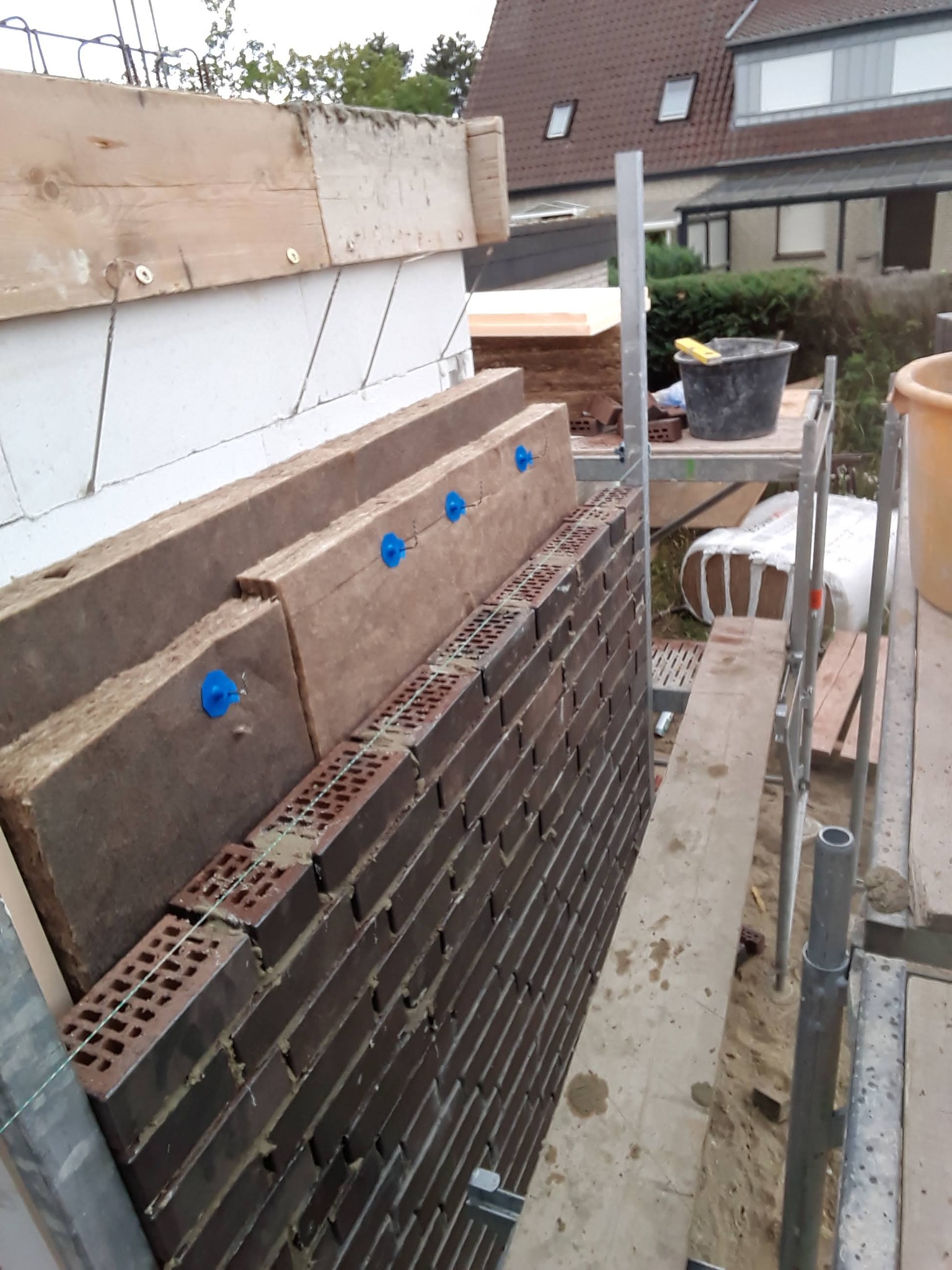 Wznoszenie ściany z cegły klinkierowej z izolacją termiczną, widoczne fragmenty rusztowania i narzędzia murarskie.