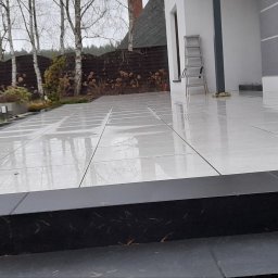 Kam-Bruk Paweł Kaczmarek - Wyłożony jasnymi płytkami taras z czarnymi obrzeżami i schodami, widoczne odbicia drzew i nieba, drabina oparta o ścianę budynku, w tle ogród z drzewami i krzewami.