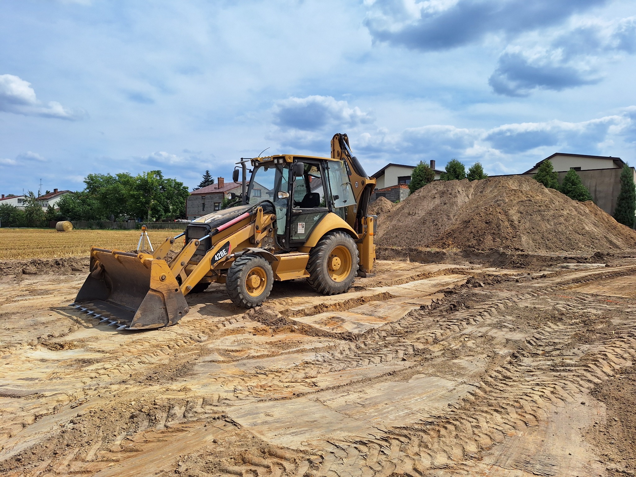 Żółta koparko-ładowarka CAT 428E na placu budowy z pryzmą ziemi w tle, widoczne ślady opon na ubitej ziemi.
