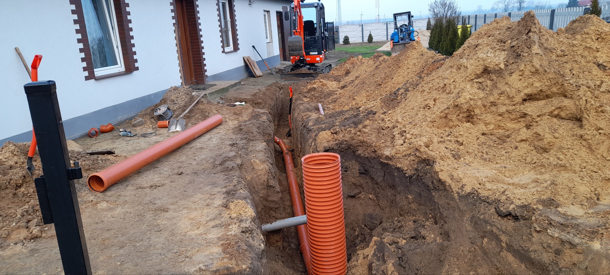 Wykop pod rury kanalizacyjne na posesji, widoczne rury z tworzywa sztucznego, mini koparka i hałda ziemi.