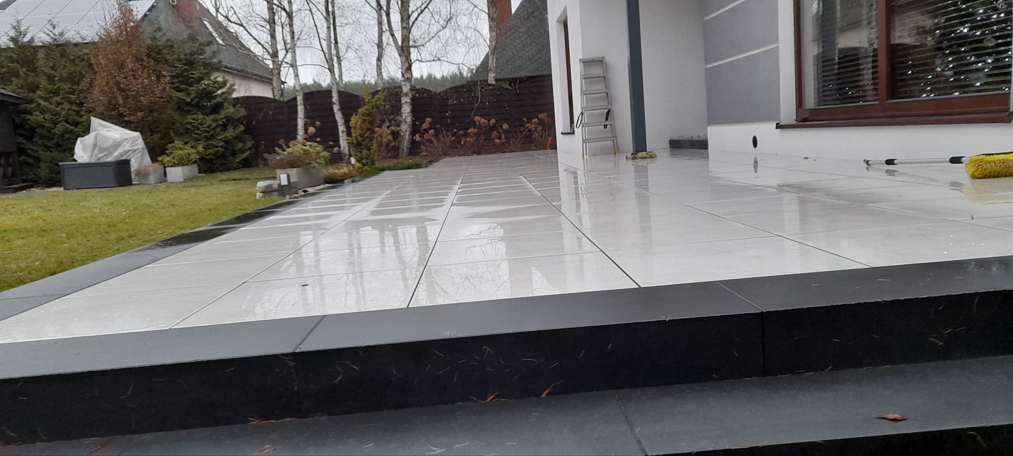 Wyłożony jasnymi płytkami taras z czarnymi obrzeżami i schodami, widoczne odbicia drzew i nieba, drabina oparta o ścianę budynku, w tle ogród z drzewami i krzewami.