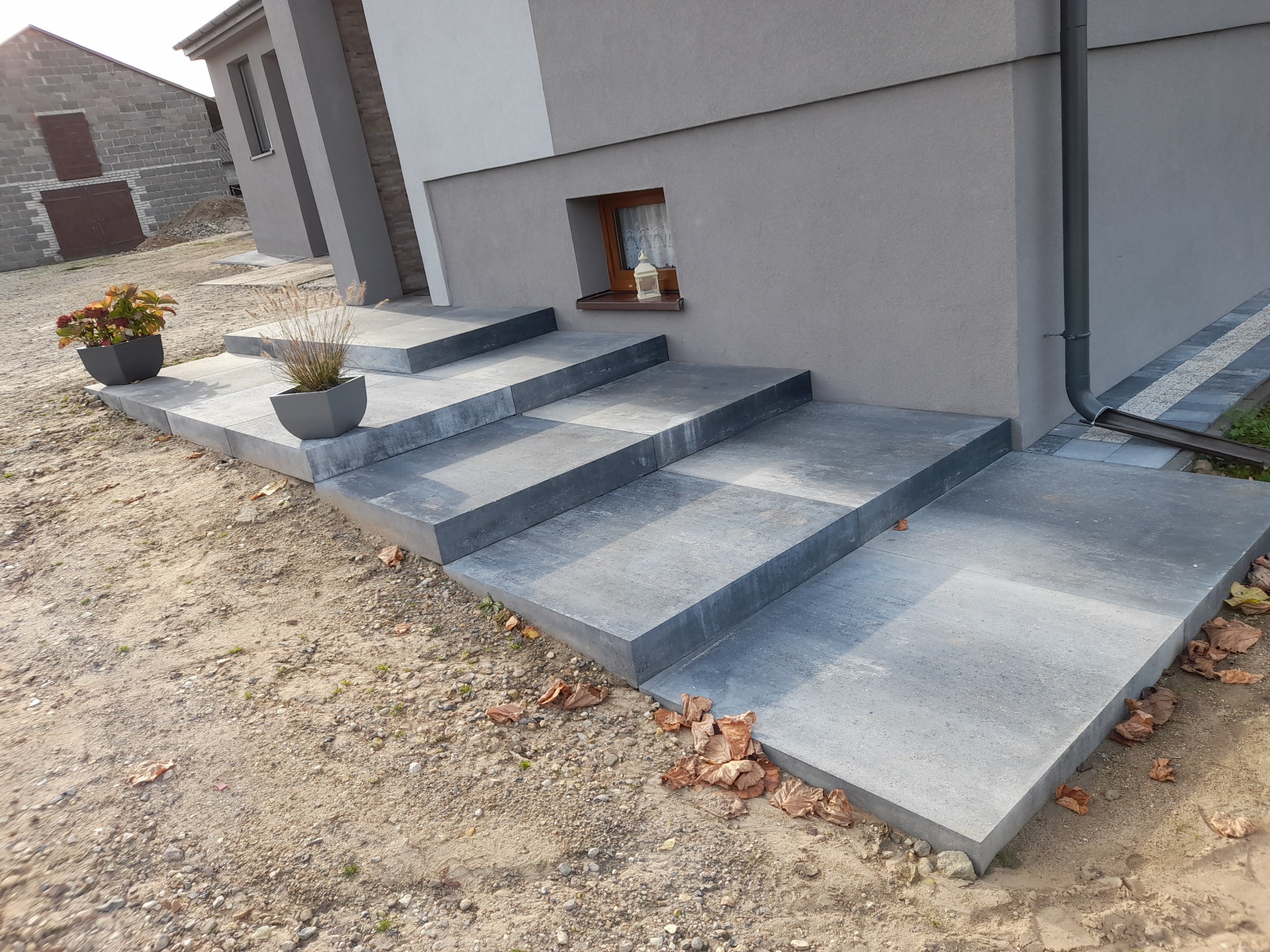 Schody wejściowe do nowoczesnego domu wyłożone szarą kostką brukową, z dekoracyjnymi donicami z roślinami po bokach, na tle surowego krajobrazu.