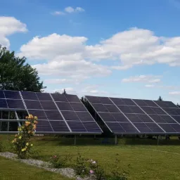 Dwa rzędy paneli słonecznych na metalowej konstrukcji, ustawione na trawniku przed budynkiem gospodarczym i drzewami, z dekoracyjnymi r&oacute;żami przy ścieżce.