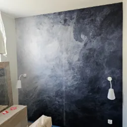 Ściana w pokoju z dekoracyjnym malowaniem imitującym chmury w odcieniach szarości i błękitu, z dwoma białymi lampkami ściennymi i zabezpieczonymi meblami.