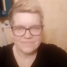 Portret kobiety w okularach, z krótkimi blond włosami, na tle beżowej ściany w łazience z widocznym fragmentem szafki.