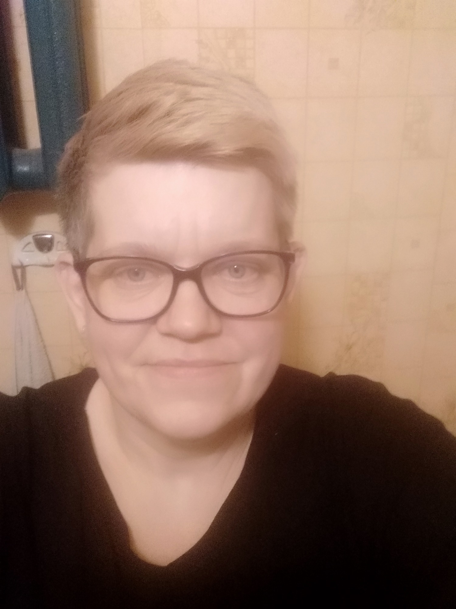 Portret kobiety w okularach, z krótkimi blond włosami, na tle beżowej ściany w łazience z widocznym fragmentem szafki.