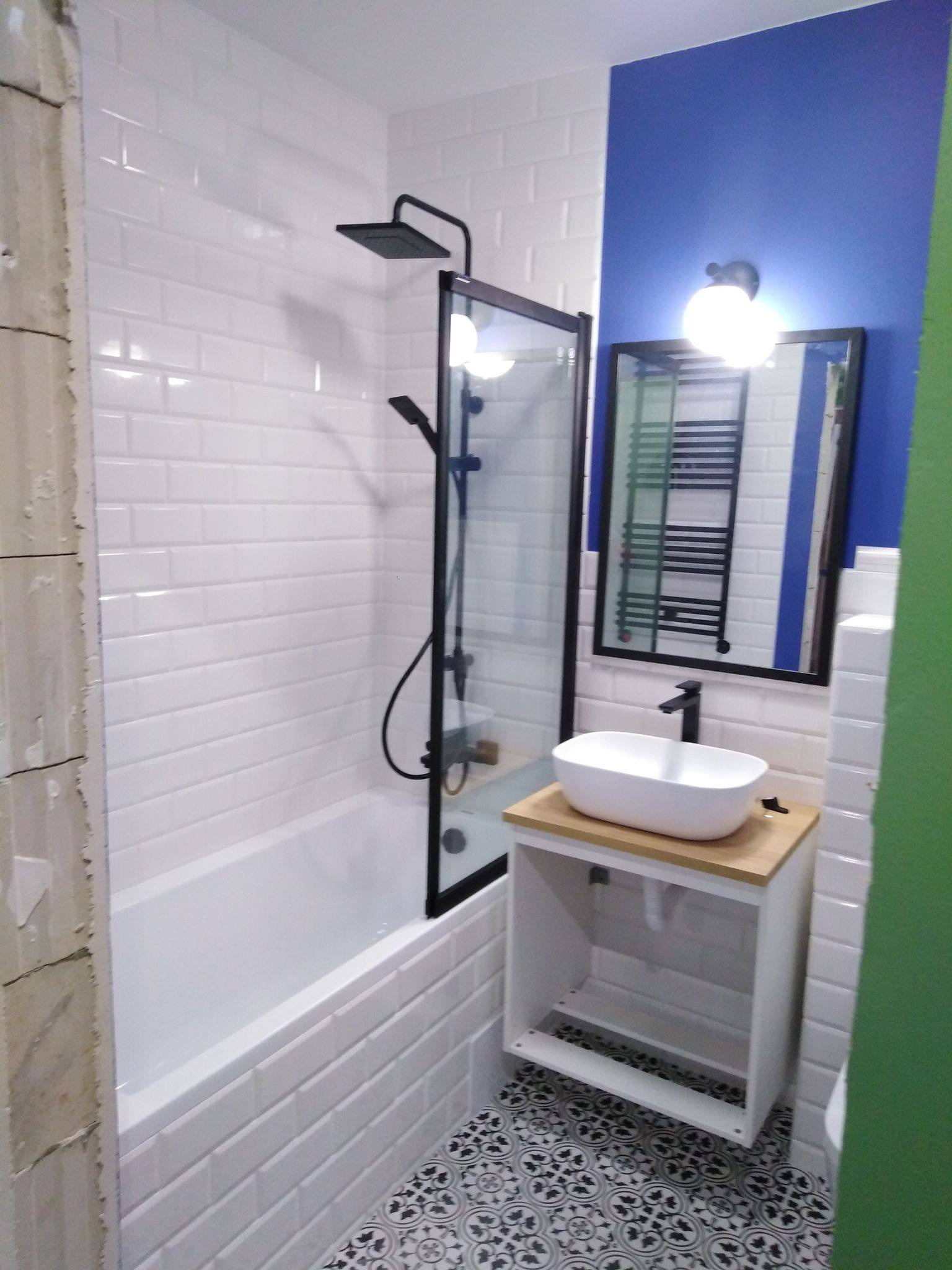 Wykończona łazienka z wanną obudowaną białymi, kafelkami typu metro, czarną armaturą, umywalką nablatową na szafce z drewnianym blatem i wzorzystą, czarno-białą podłogą; fragment ściany w kolorze...